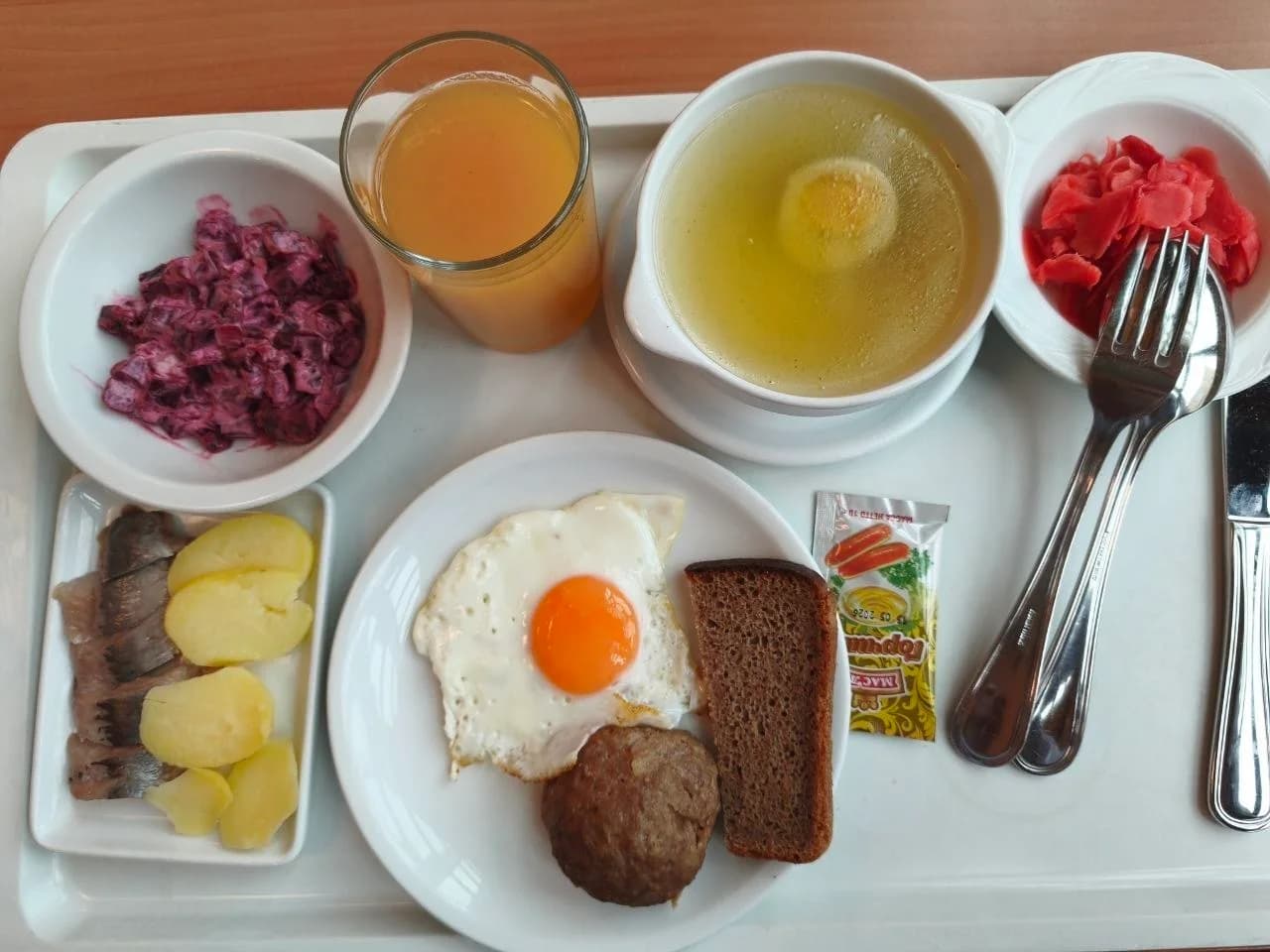 Broth with egg, a cutlet with a fried egg and rye bread, herring with boiled potatoes, beet salad, pickled vegetables, and juice.